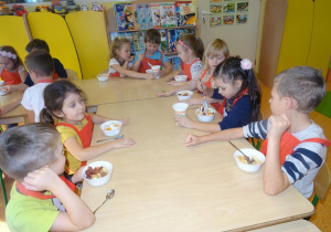 Grupa dzieci siedzi przy stole z wypełnionymi owocami miseczkami. Dwoje dzieci nakłada jogurt.